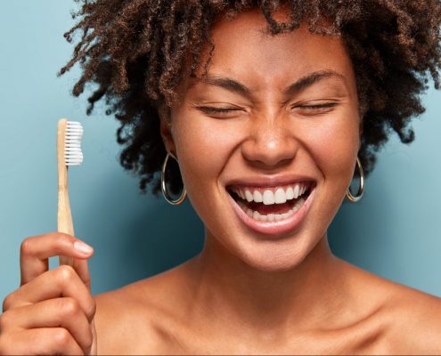 oral microbiome smiling with curly hair, laughs while has morning routines, shows bright smile, holds toothbrush, stands with bare shoulders over blue background