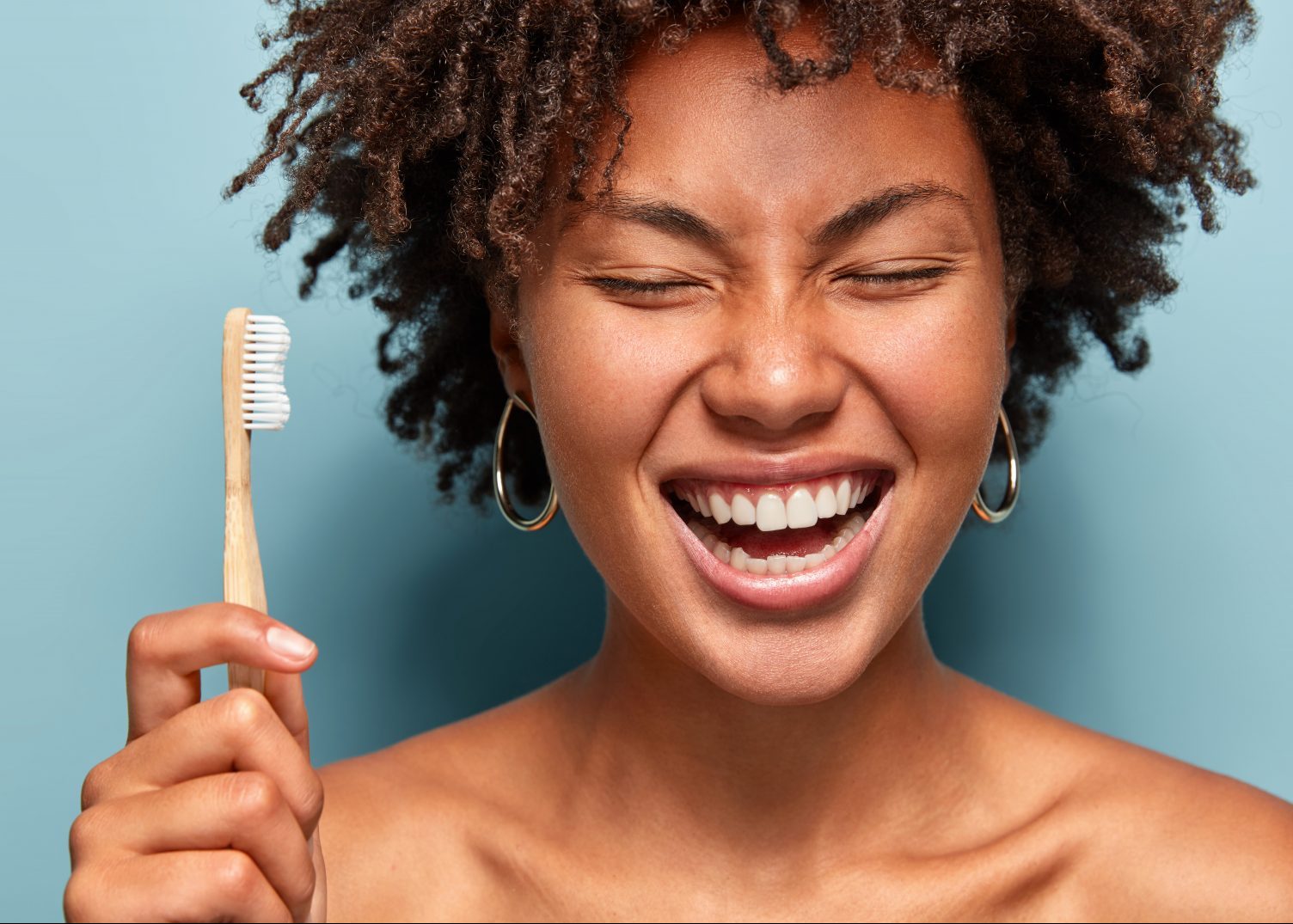 oral microbiome smiling with curly hair, laughs while has morning routines, shows bright smile, holds toothbrush, stands with bare shoulders over blue background